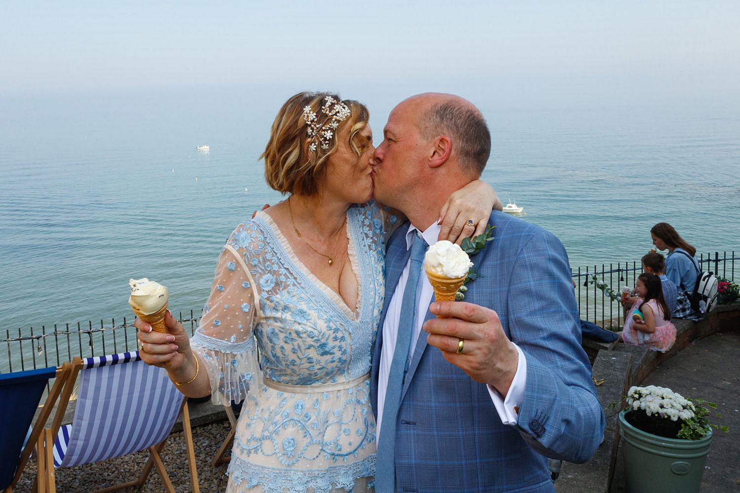 Newlywed couple kissing while holding ice cream cones at a seaside cliff with ocean views on their wedding day.