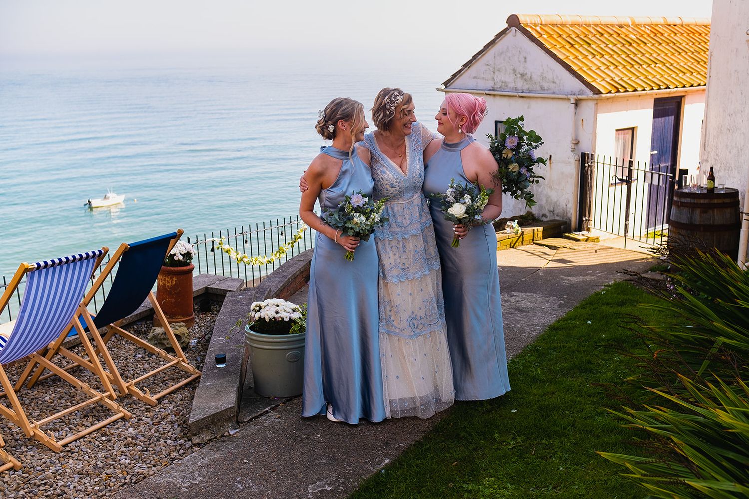 Three bridesmaids in blue dresses stand together by the seaside, holding bouquets near a charming coastal cottage.