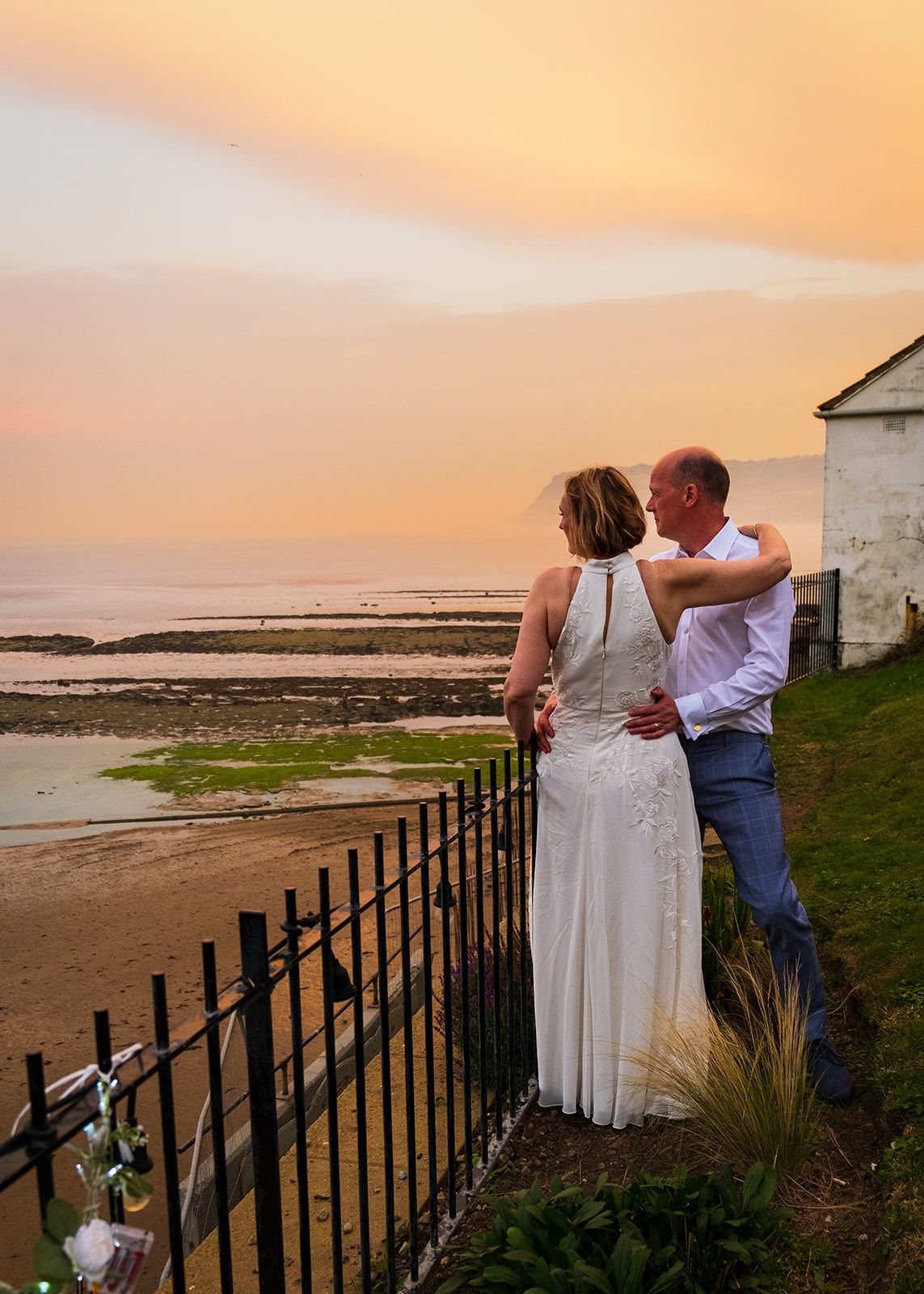 Couple embracing at sunset by the seaside, bride in white gown and groom in casual attire near iron fence.