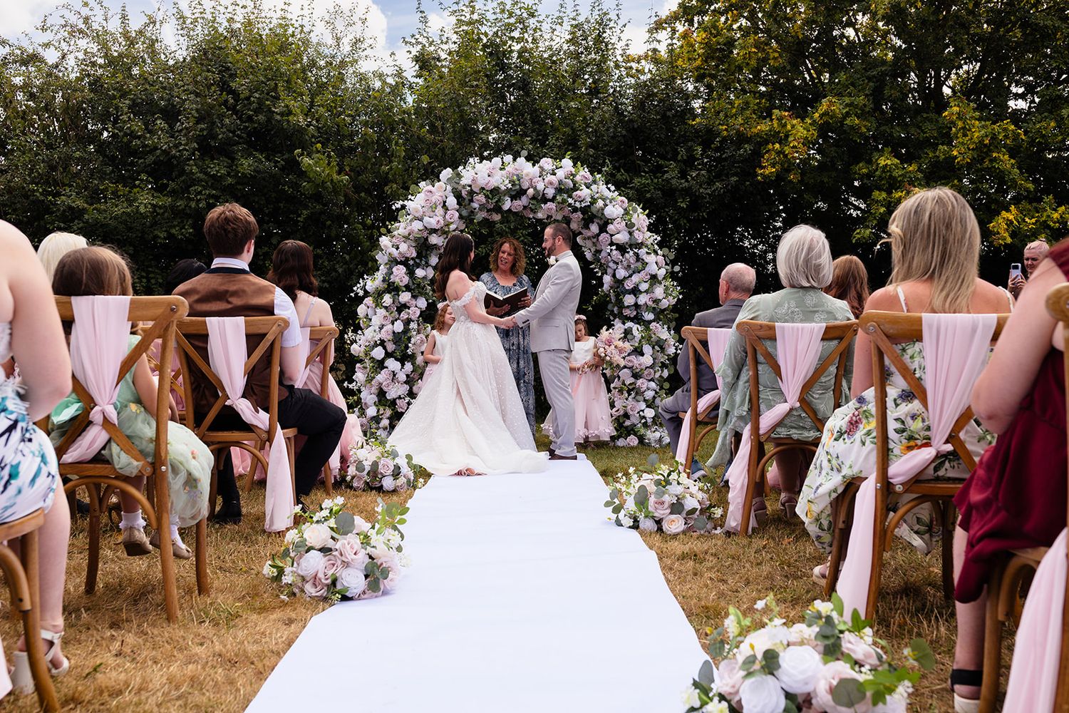 Outdoor wedding ceremony with couple at floral arch altar, guests seated on wooden chairs along white aisle runner.