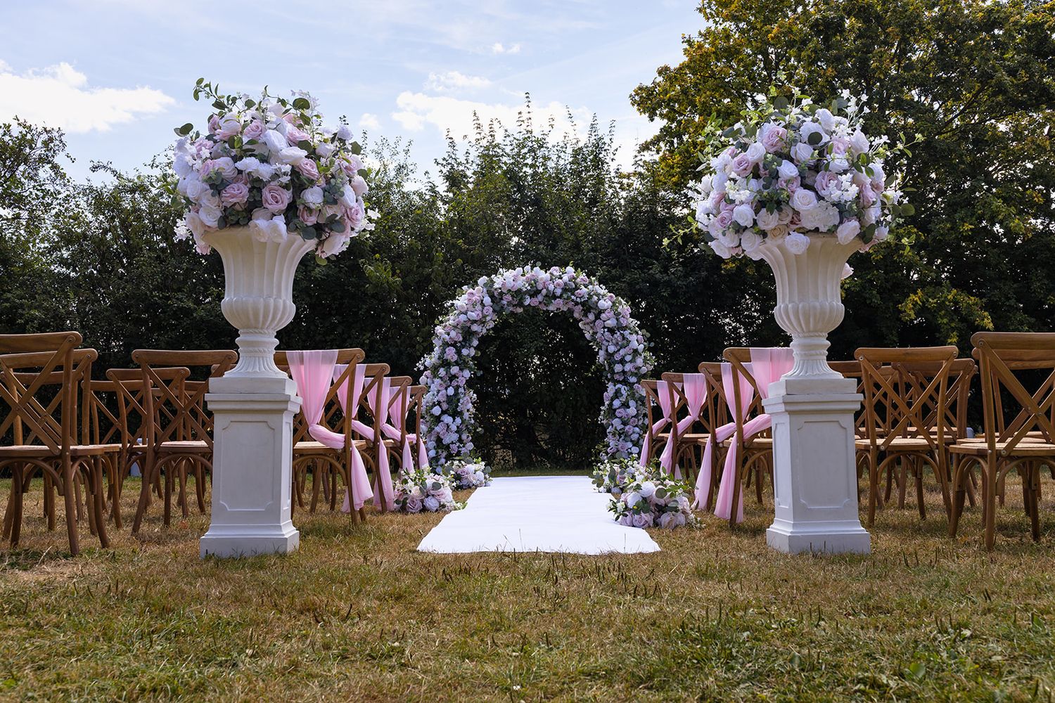 Outdoor wedding ceremony setup with floral arch, white pedestals, and wooden chairs on green lawn.