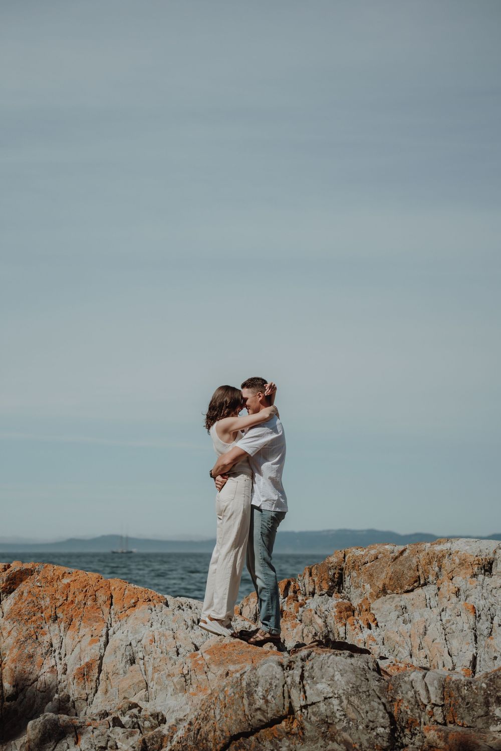 Couple embracing and kissing on rocky coastal cliffs with ocean horizon in background. Engagement photoshoot. Victoria, BC, Vancouver Island