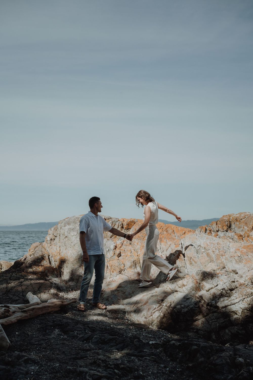 Couple holding hands on scenic rocky coastline with driftwood and ocean in background. Engagement photoshoot. Victoria, BC, Vancouver Island