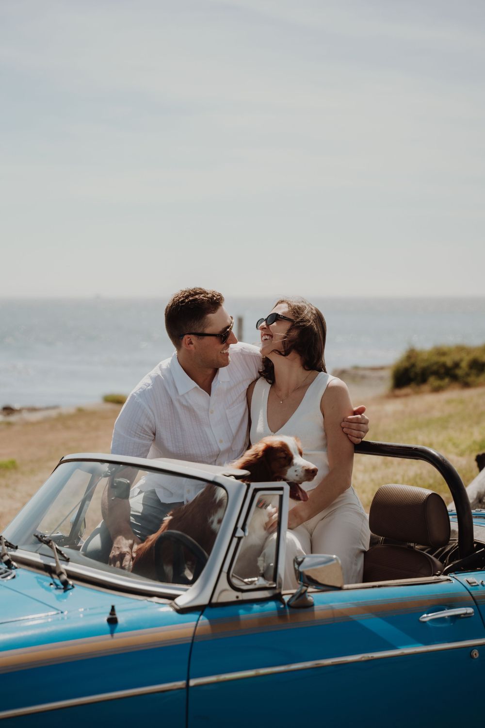 Stylish couple with dog sitting in vintage blue convertible car near oceanside meadow. Engagement photoshoot. Victoria, BC, Vancouver Island