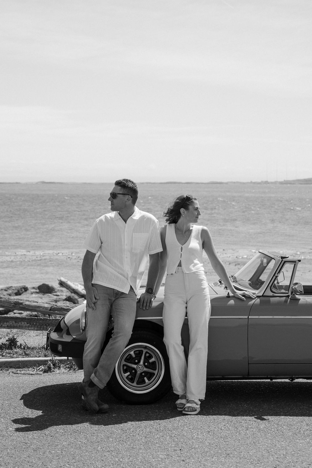 Black and white photo of a couple leaning against a classic convertible car by the sea.  Engagement photoshoot. Victoria, BC, Vancouver Isla