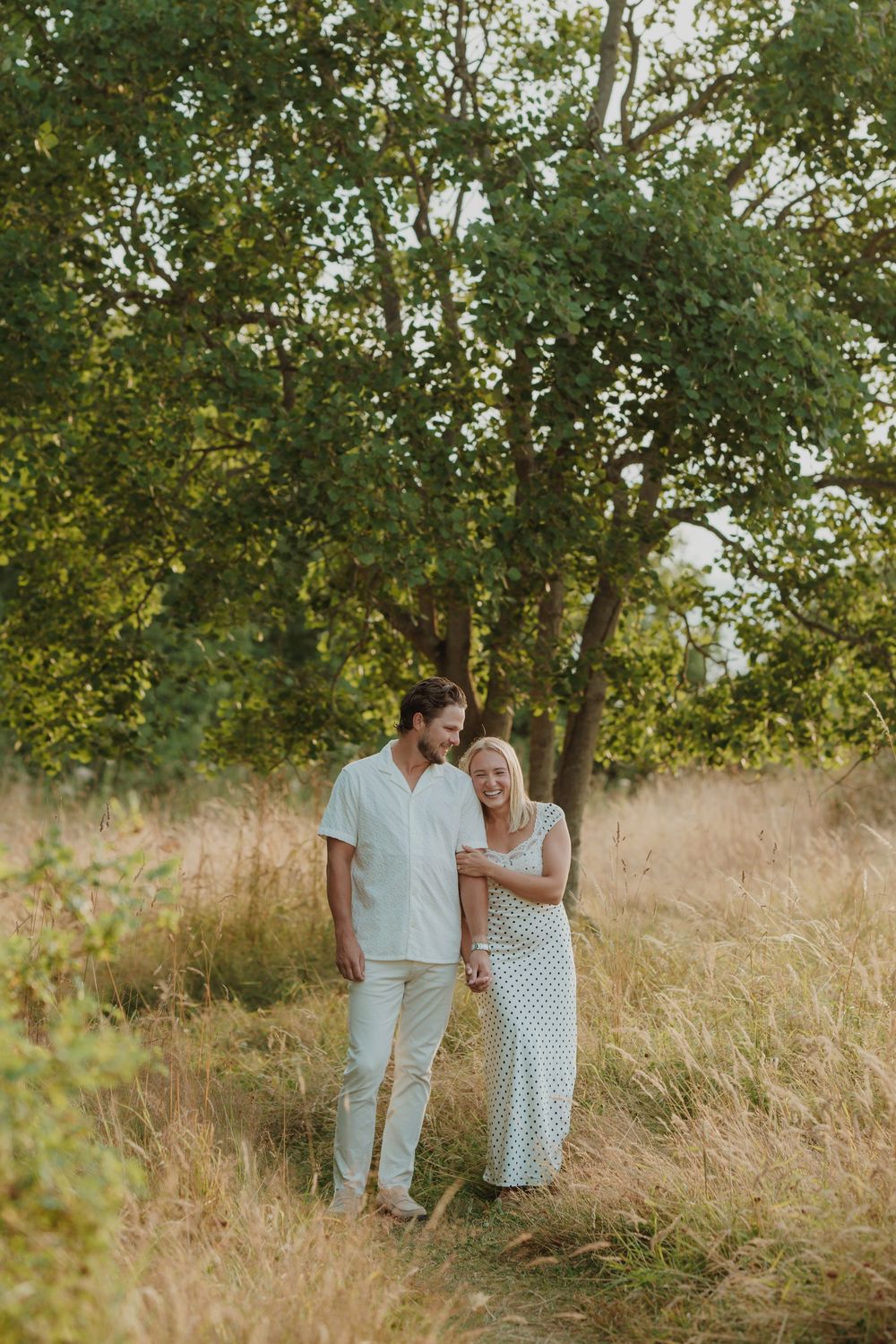 A couple walking hand in hand through a golden meadow surrounded by lush green trees. Engagement photoshoot. Victoria, BC, Vancouver Island.