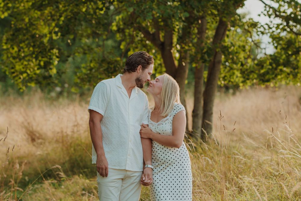 Couple sharing a tender moment in a golden meadow with lush green trees in warm sunlight. Engagement photoshoot. Victoria, BC.