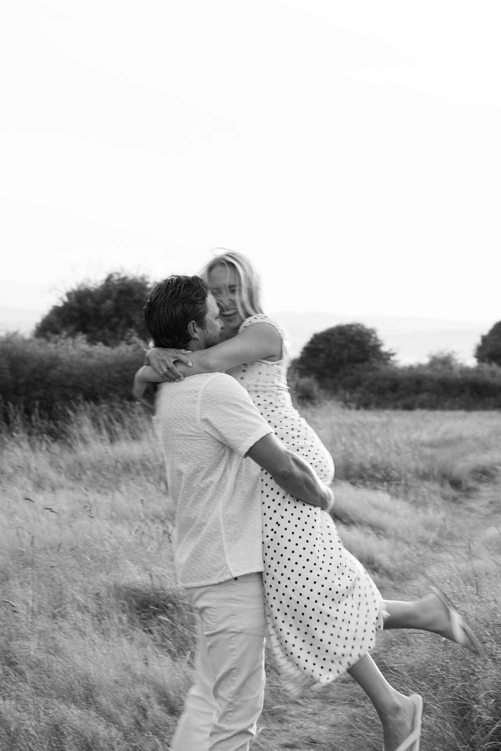 Black and white photo of a man lifting a woman in a joyful embrace in a field. Engagement photoshoot. Victoria, BC, Vancouver Island.