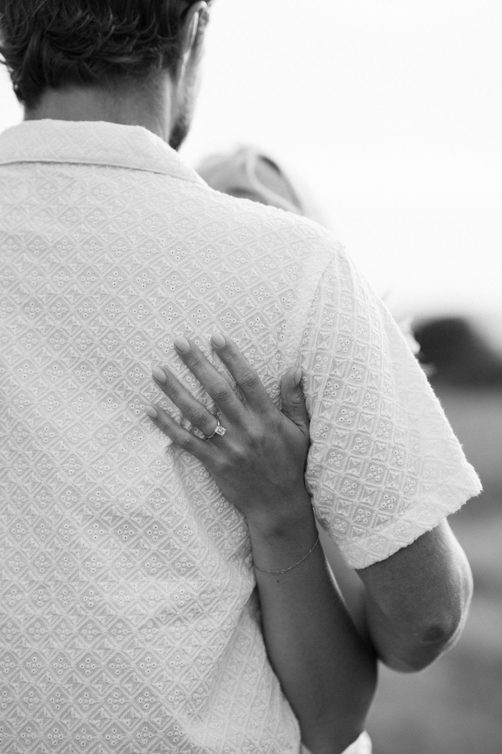Black and white close-up of a woman placing her hand on a mans back showing her ring. Engagement photoshoot. Victoria, BC, Vancouver Island.