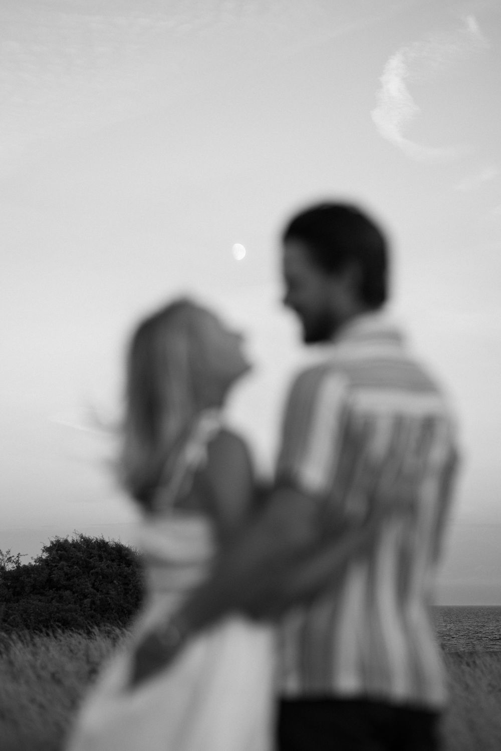 Blurry black and white photo of a couple embracing in a field under a crescent moon at dusk. Engagement photoshoot. Victoria, BC.