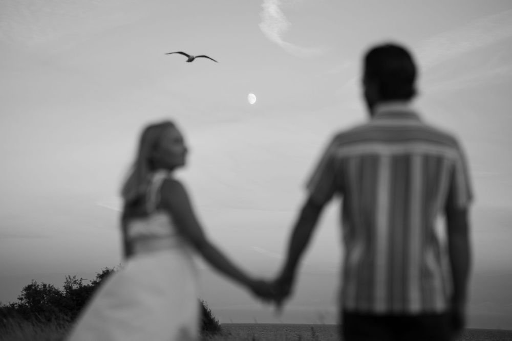 Black and white photo of couple holding hands watching a bird fly under a crescent moon at dusk. Engagement photoshoot. Victoria, BC.