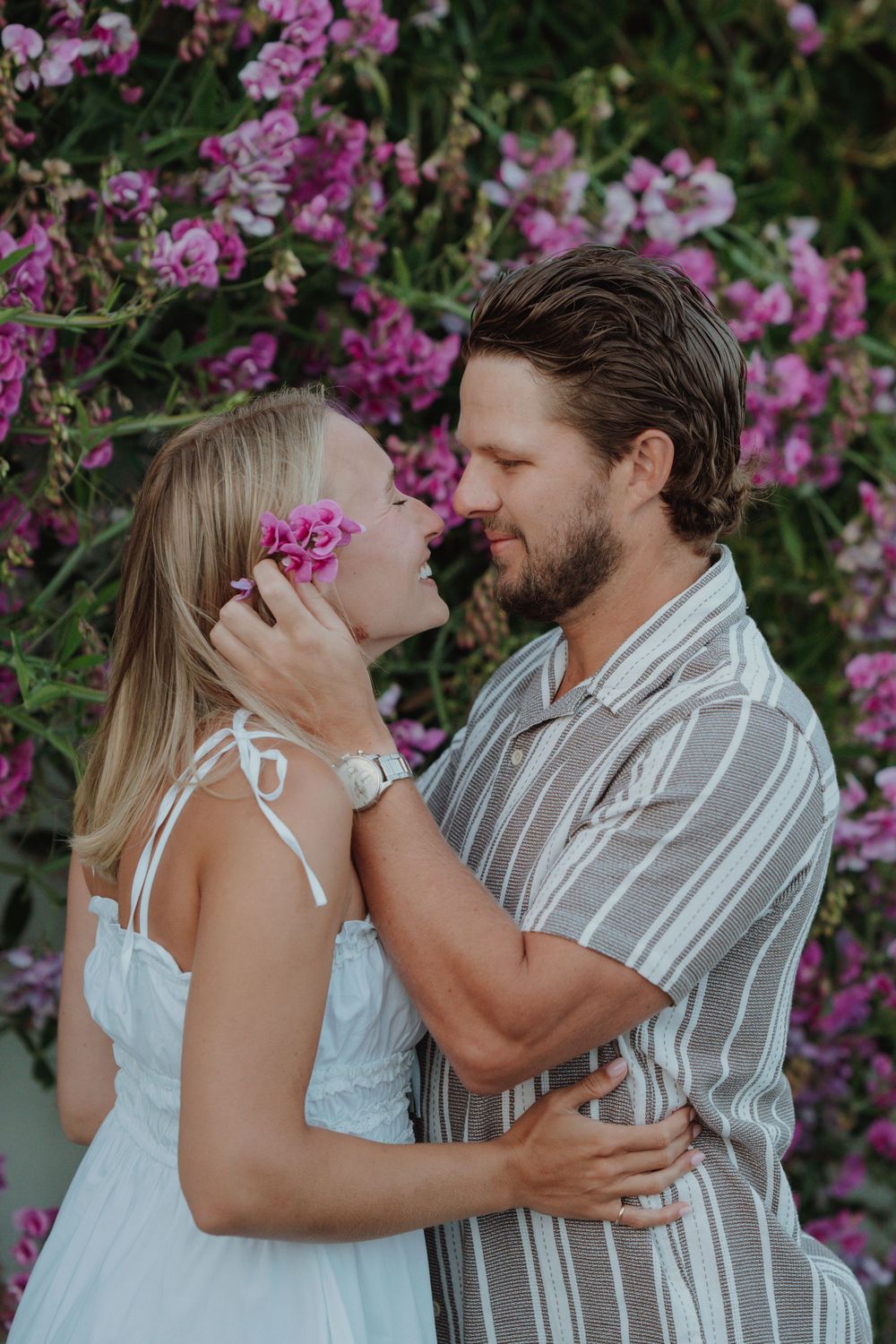 Romantic couple kissing amid pink flowers, woman in white dress holding a bloom, man in striped shirt. Engagement photoshoot. Victoria BC