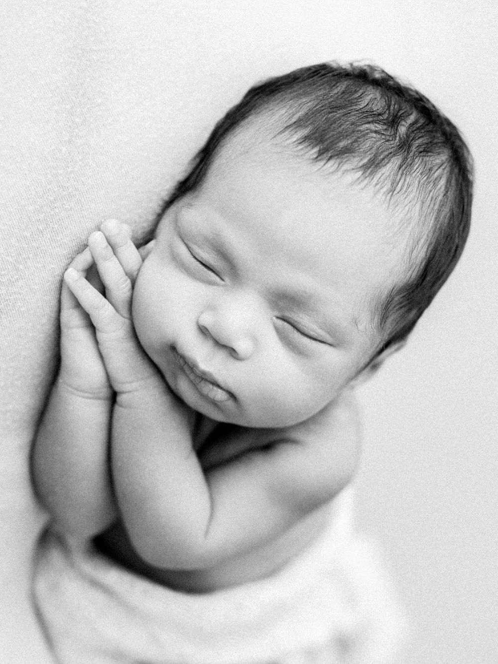 Sweet black and white portrait of a sleeping newborn baby with hands gently resting against their cheek.