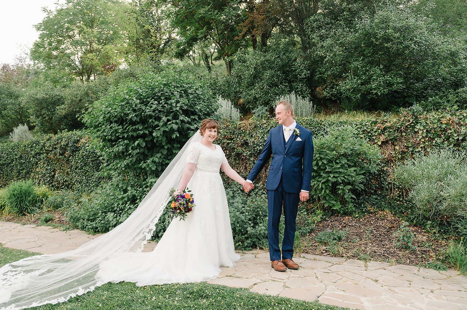 Wedding couple poses on garden path with flowing white dress and veil against lush greenery background.