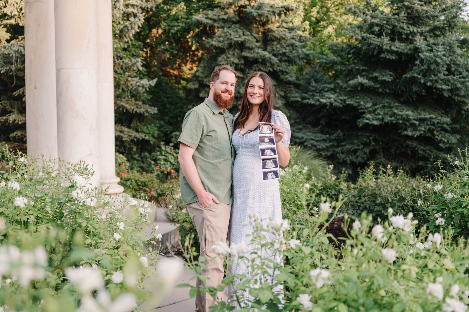 Couple standing among blooming flowers with white columns in background.
