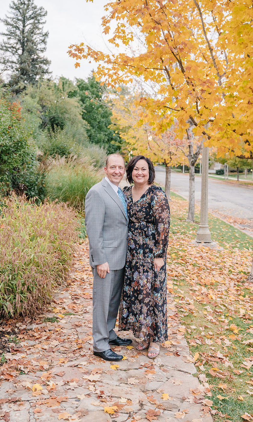 Couple in formal wear posing on leaf-covered path with golden autumn trees.
