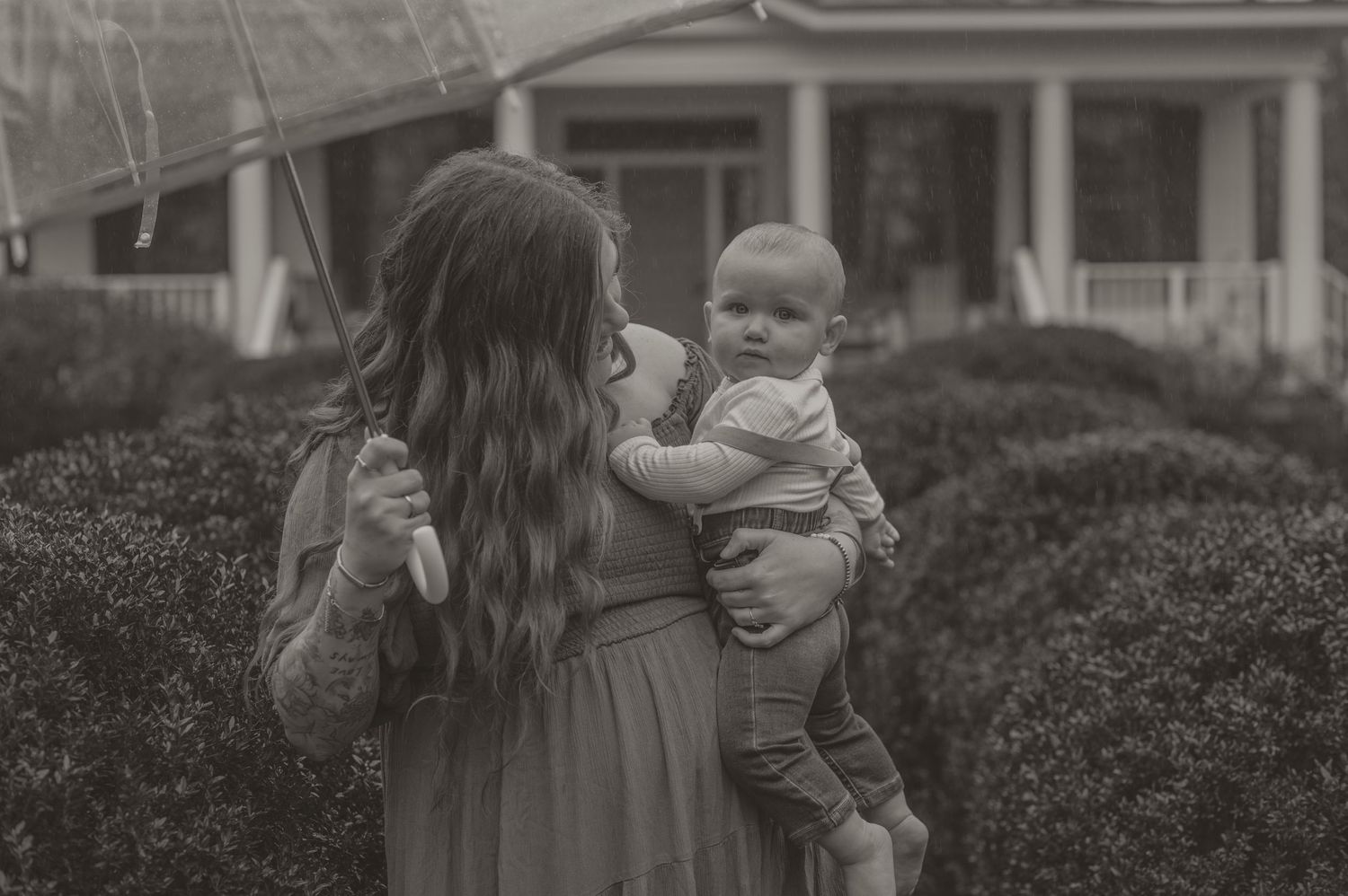 A black and white portrait of a mom holding a baby outdoors in front of bushes and a house. photo by Samantha Campbell imagery