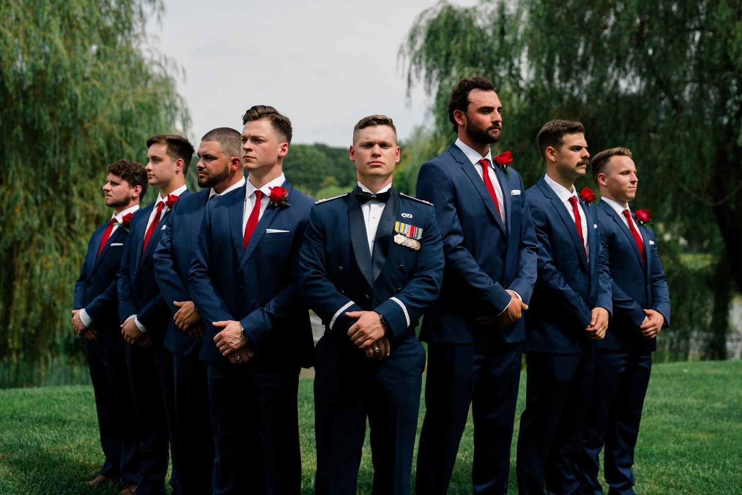Groomsmen in navy suits and red ties pose together outdoors under willow trees.