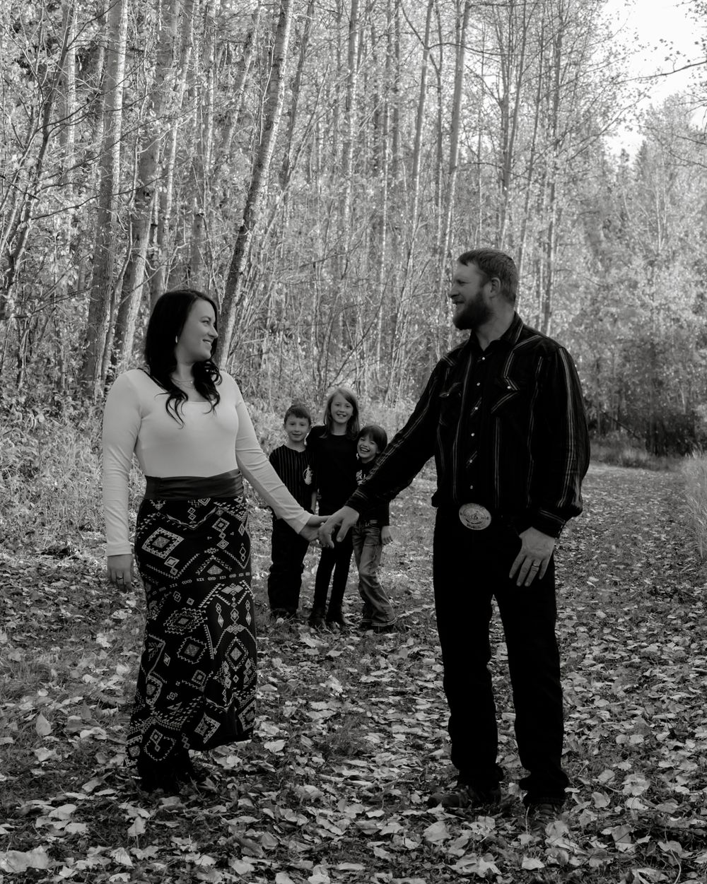 Black and white photo of a couple holding hands on a leaf-covered forest path while three children stand behind them.