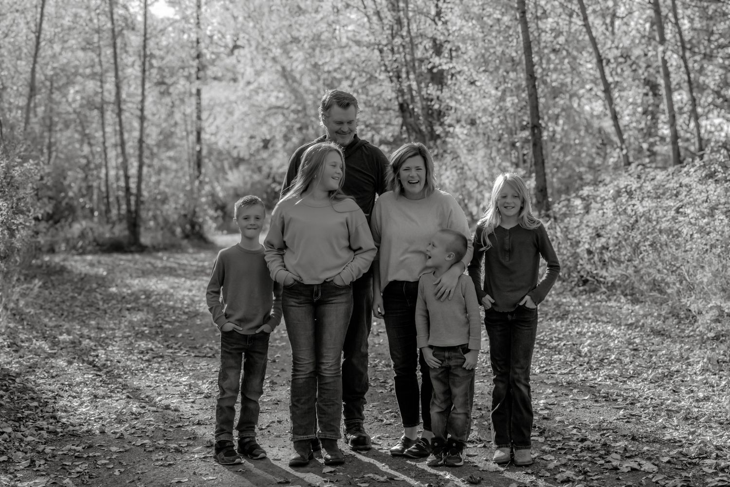 Black and white family portrait of six people posing together on a leaf-covered forest path in autumn.