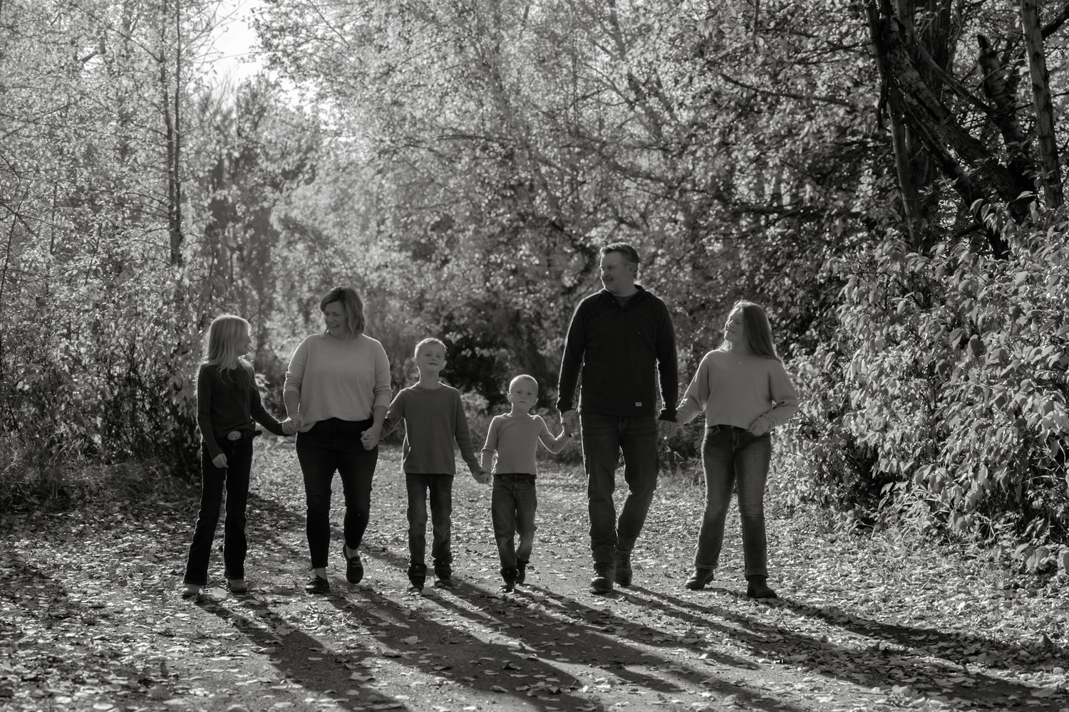 Black and white photo of a family of six holding hands walking down a sunlit autumn forest path.
