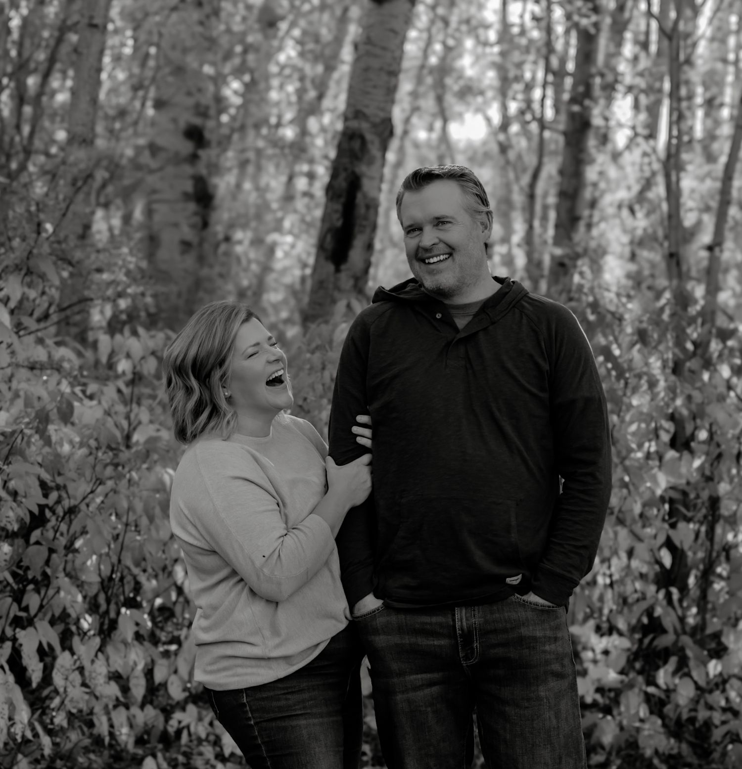 Black and white photo of a laughing couple standing together among birch trees in a forest setting.