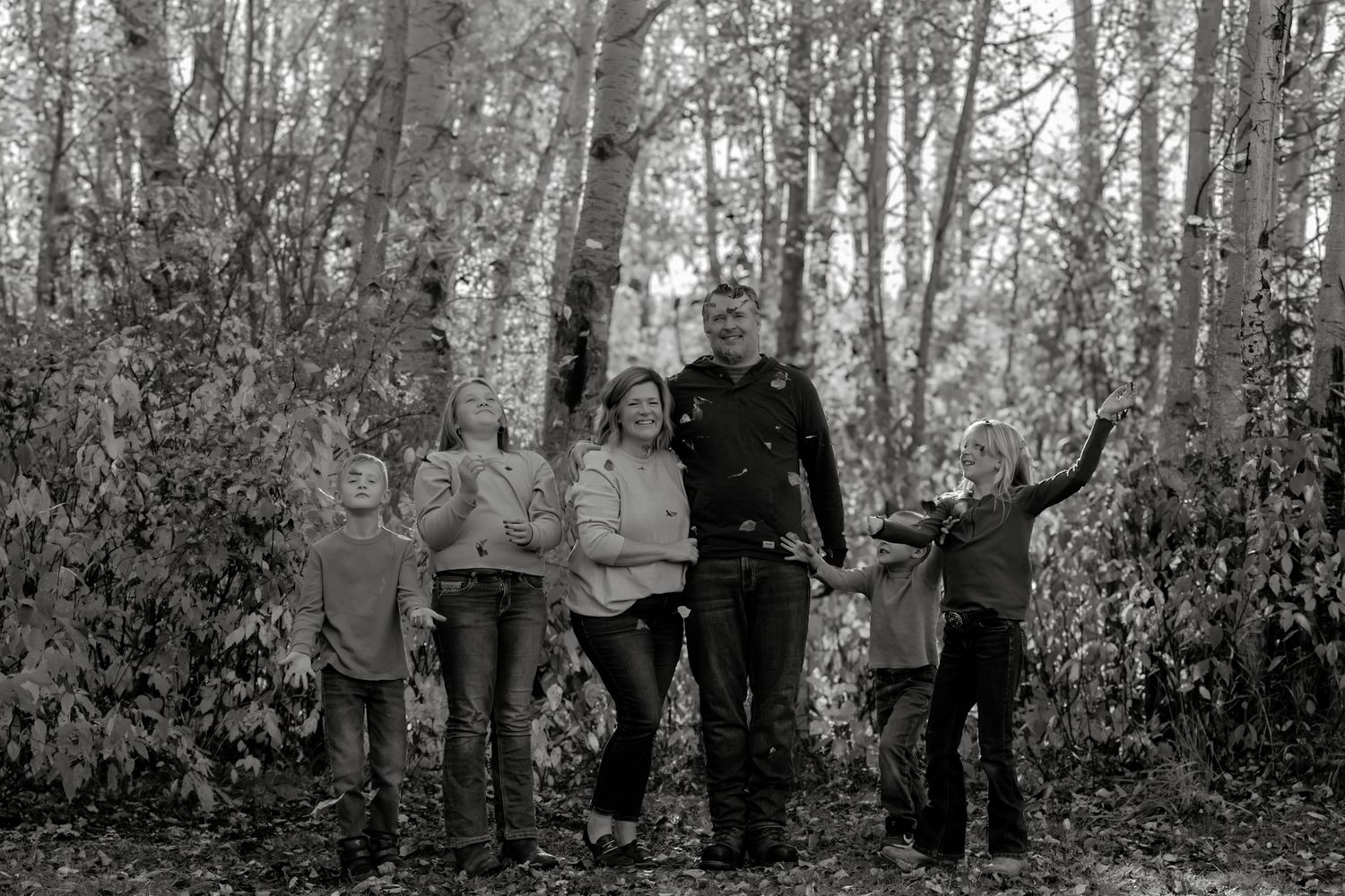 Black and white photo of a playful family of six posing among tall birch trees in a forest.