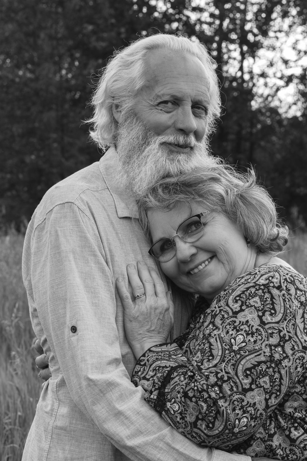 Black and white portrait of a smiling elderly couple embracing outdoors, man with white beard hugging woman with glasses.