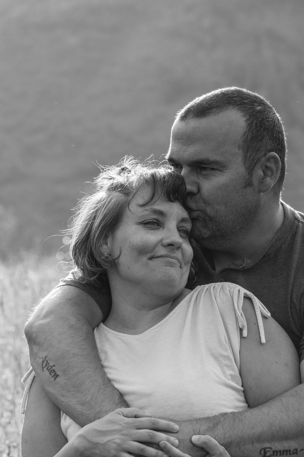 Black and white photo of a man kissing a smiling woman on the cheek while embracing her outdoors.