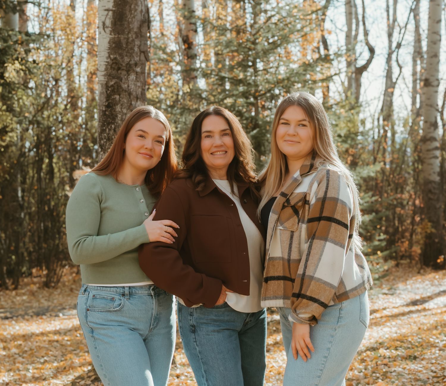 Three women smile together in a sunny autumn forest with golden leaves and birch trees behind them.
