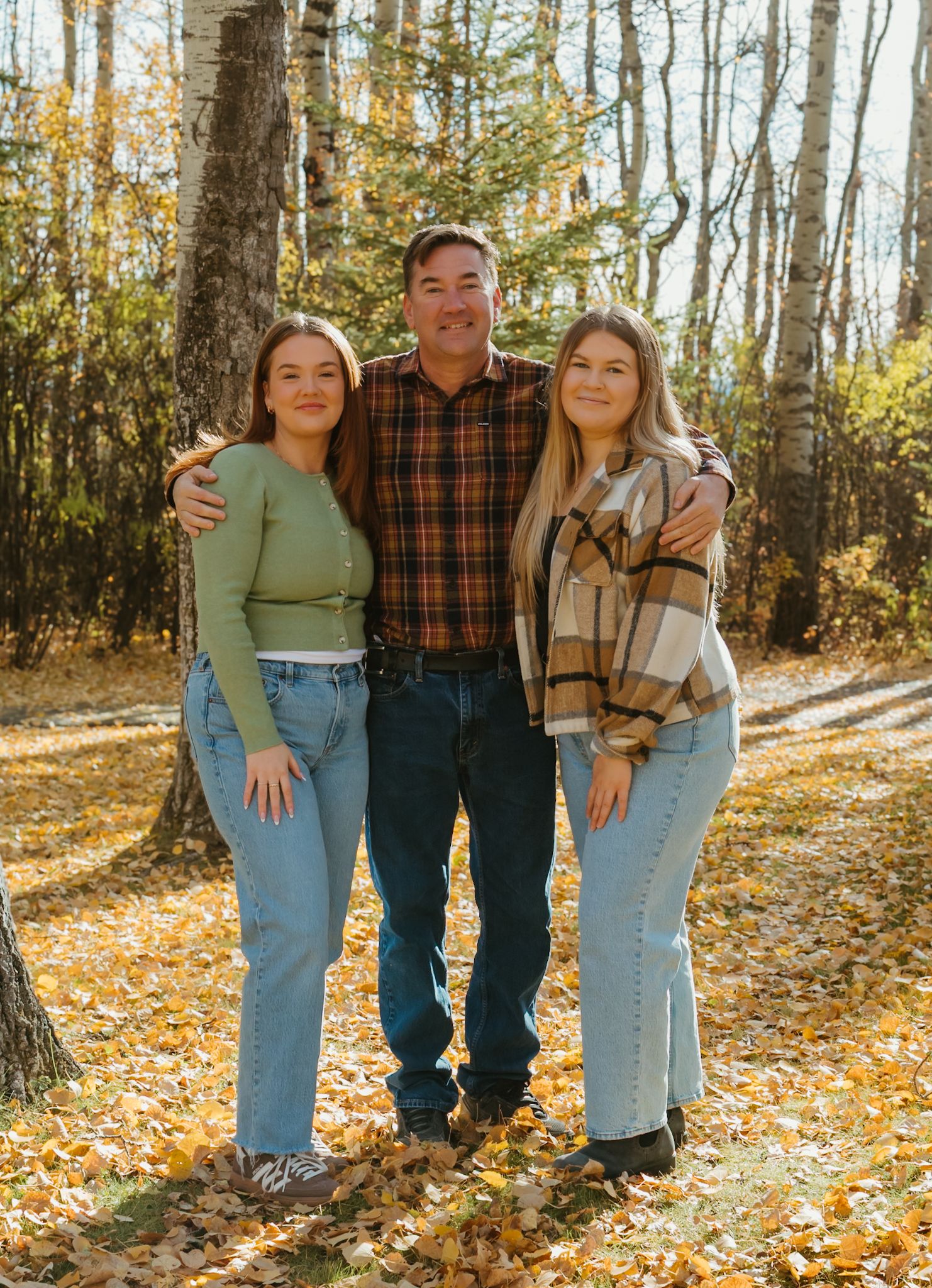 A man smiles with two young women on either side in a colorful autumn forest setting.