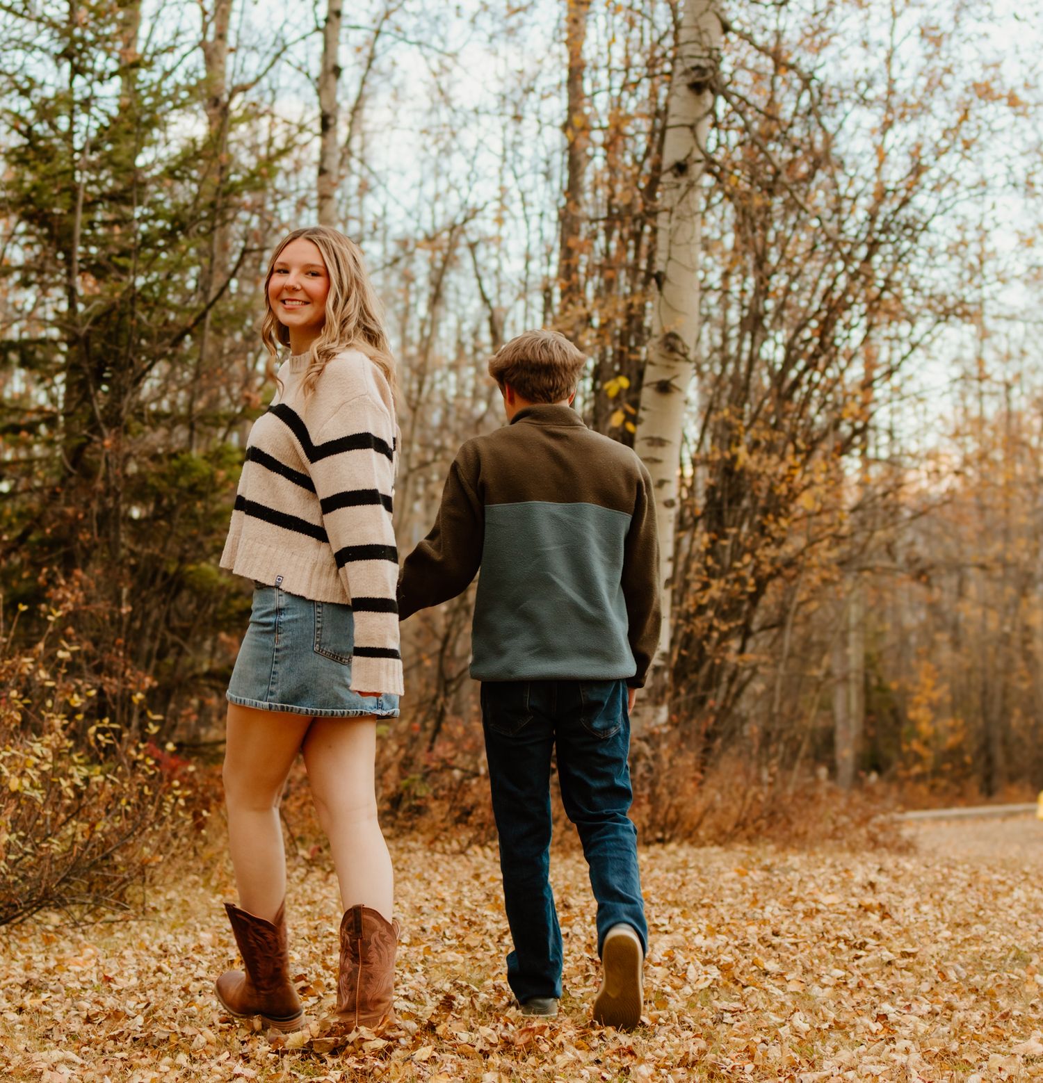 A smiling young woman in a striped sweater and boots walks hand in hand with a man through autumn leaves.