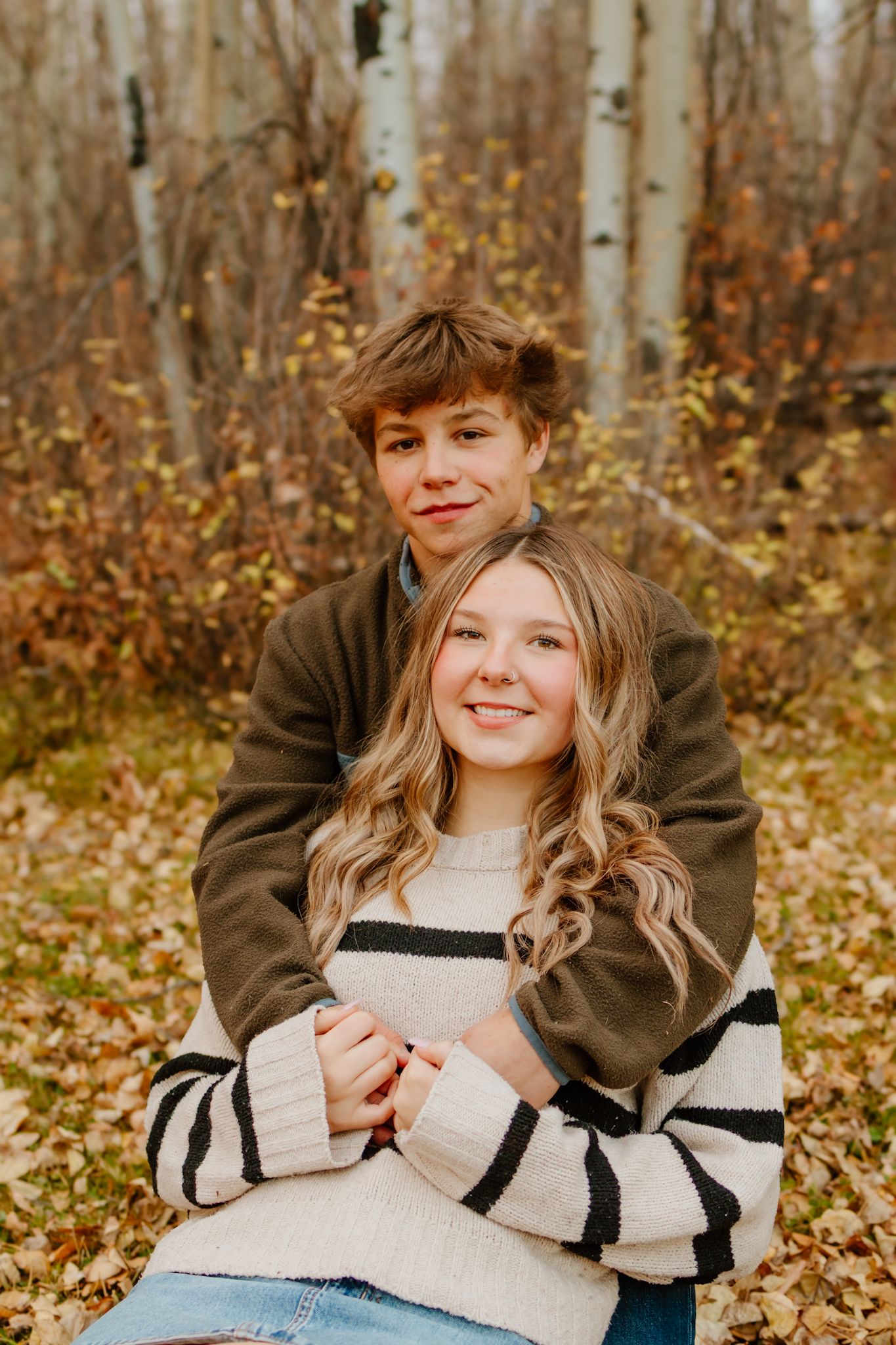 Two smiling teens embrace in autumn forest, girl in striped sweater, boy in olive fleece.