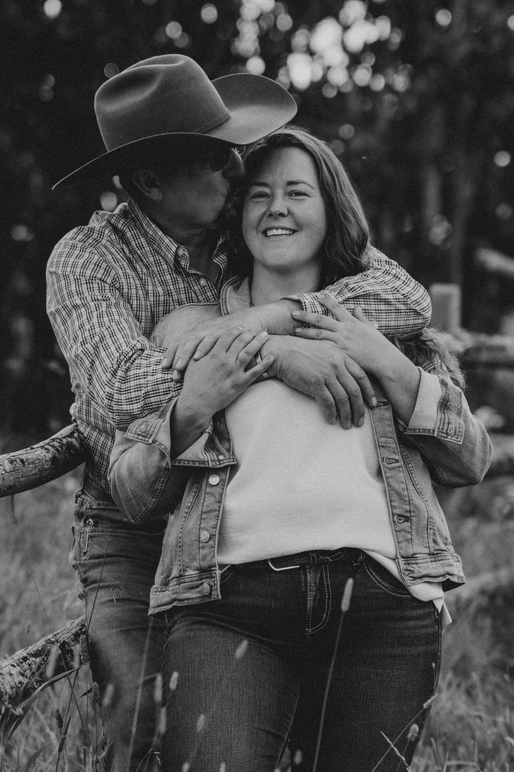 Black and white photo of a couple embracing outdoors, man in cowboy hat kissing woman's cheek as she smiles.