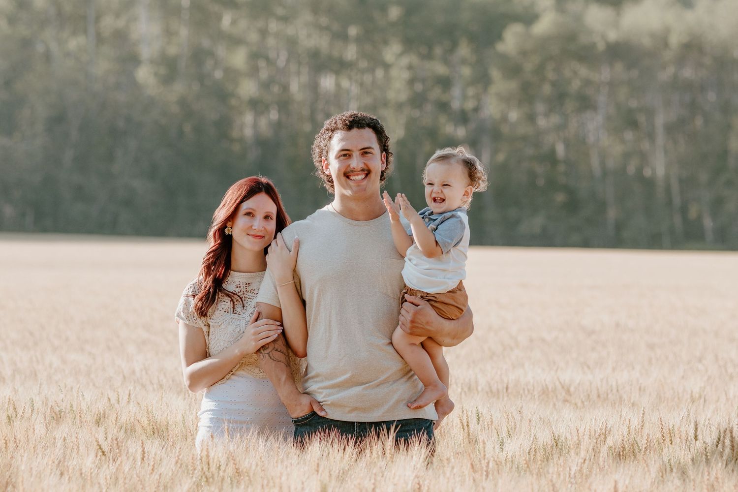 Smiling family with a laughing baby stands together in a sunlit golden wheat field.