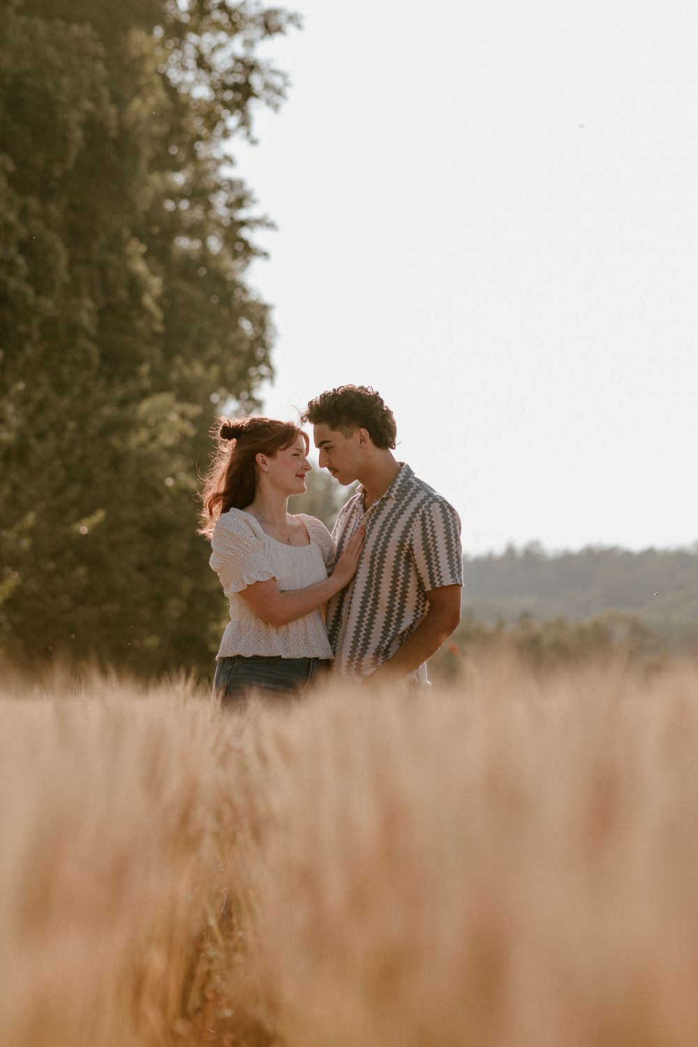 Young couple gazes at each other lovingly while standing in a golden sunlit wheat field.