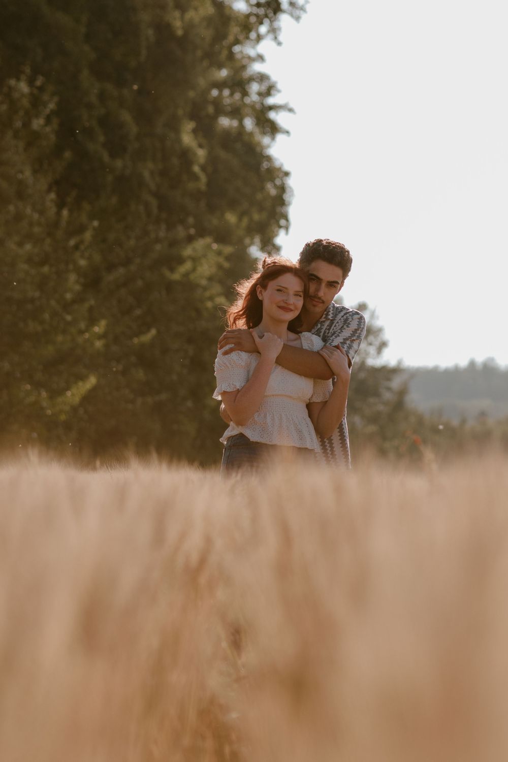 Couple embraces warmly in a golden wheat field with tall green trees in the background.
