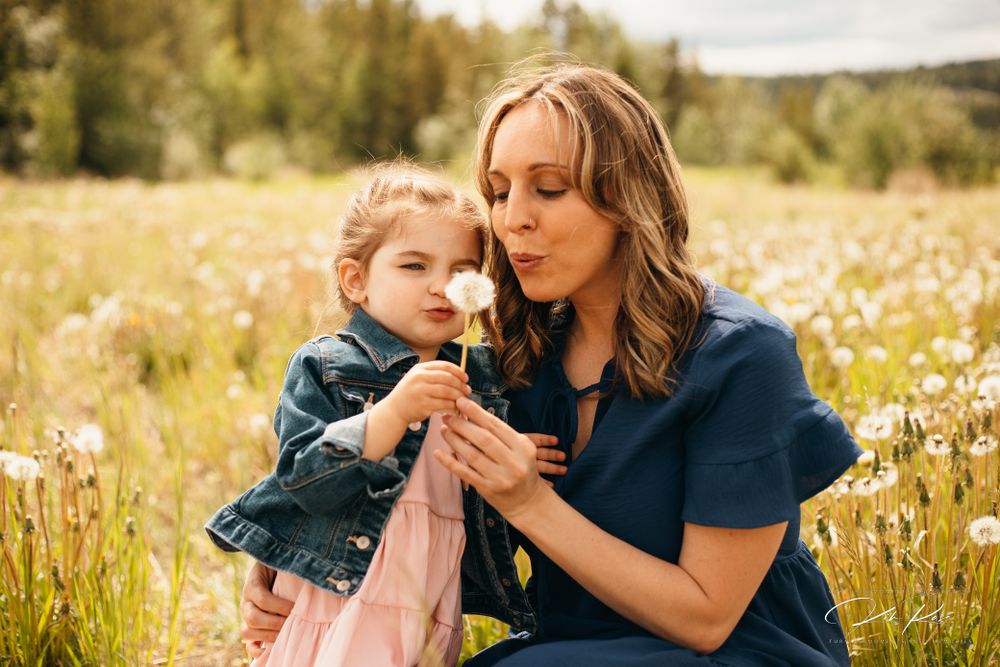 A mother and young daughter blow a dandelion together in a sunny wildflower meadow.