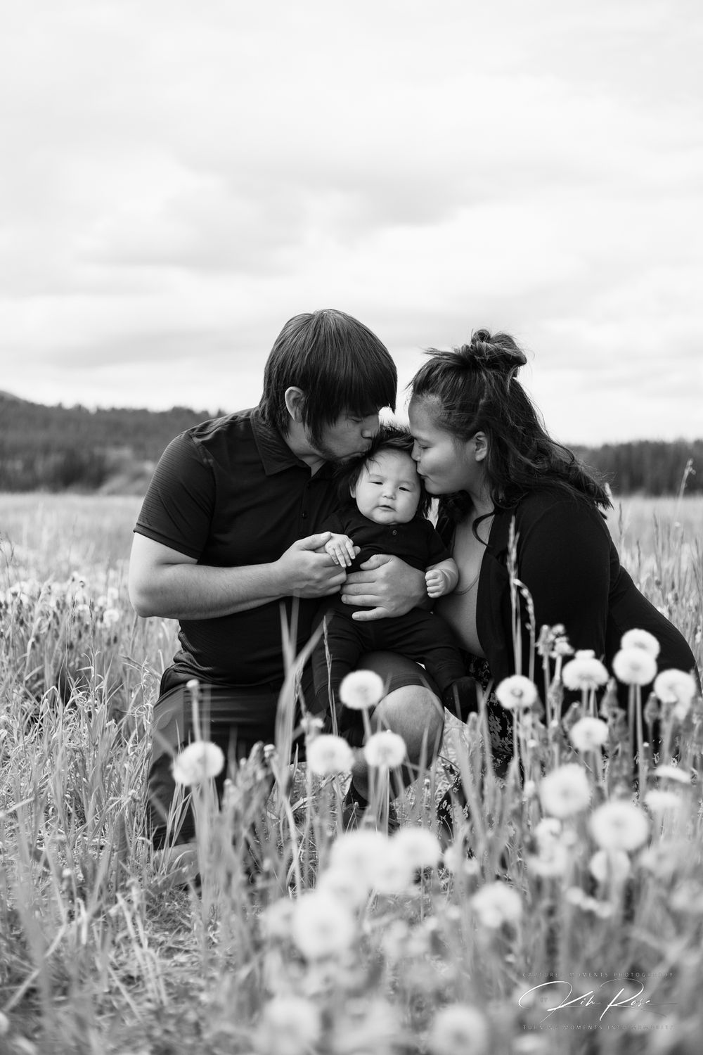 A couple kissing their infant baby on the cheeks in a dandelion field, black and white.