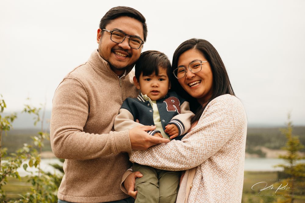 A happy Asian family of three smiling together outdoors with a scenic background.