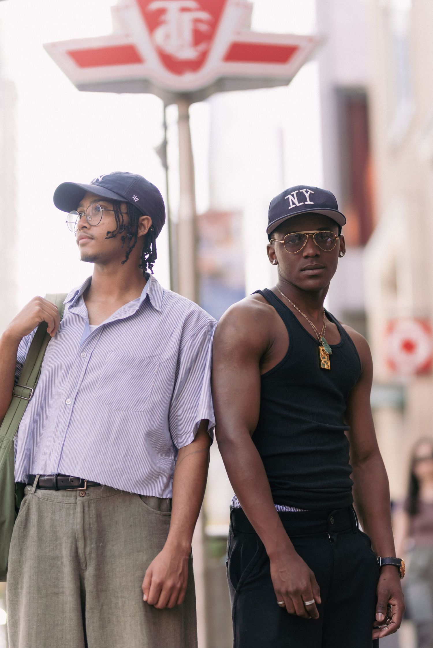 Two people standing beneath a sign, wearing NY caps in urban setting.