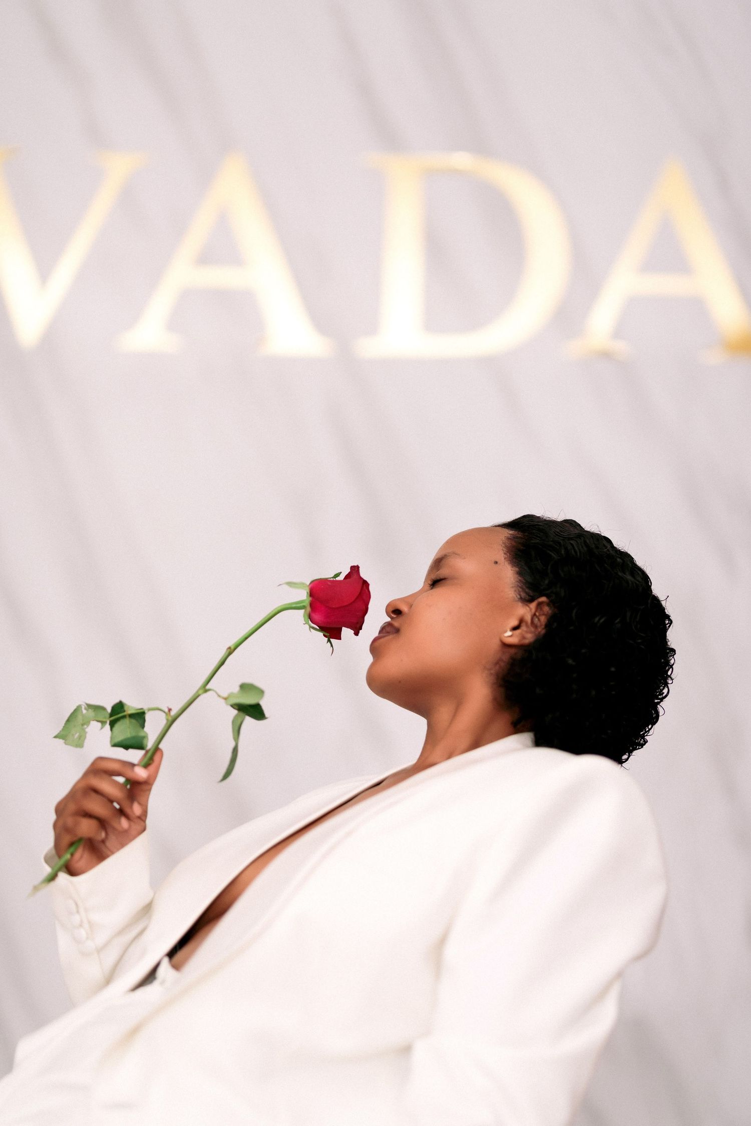 Artistic portrait in white blazer smelling red rose against VADA backdrop.