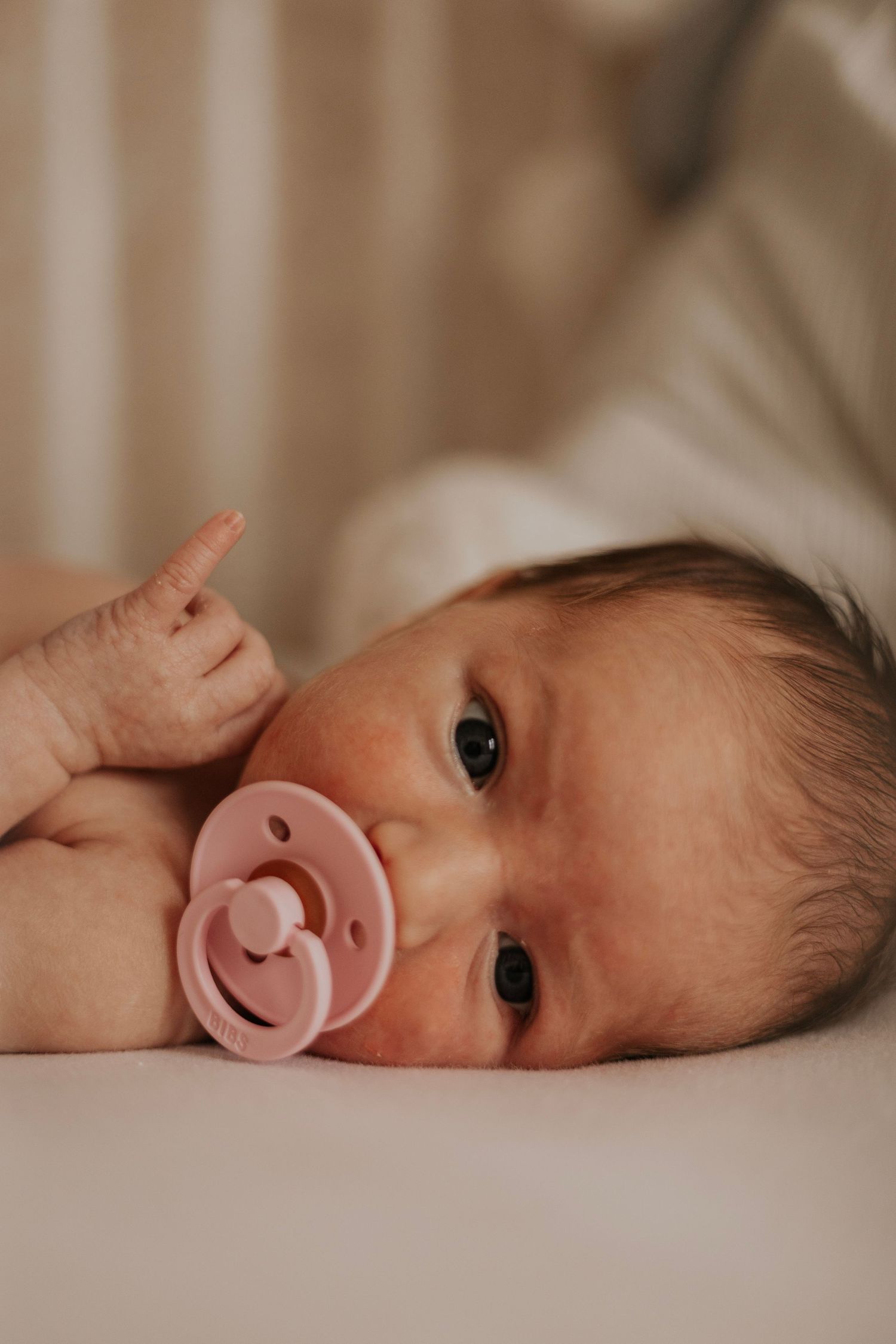 A baby lying down with a pink pacifier reaches up with one finger.
