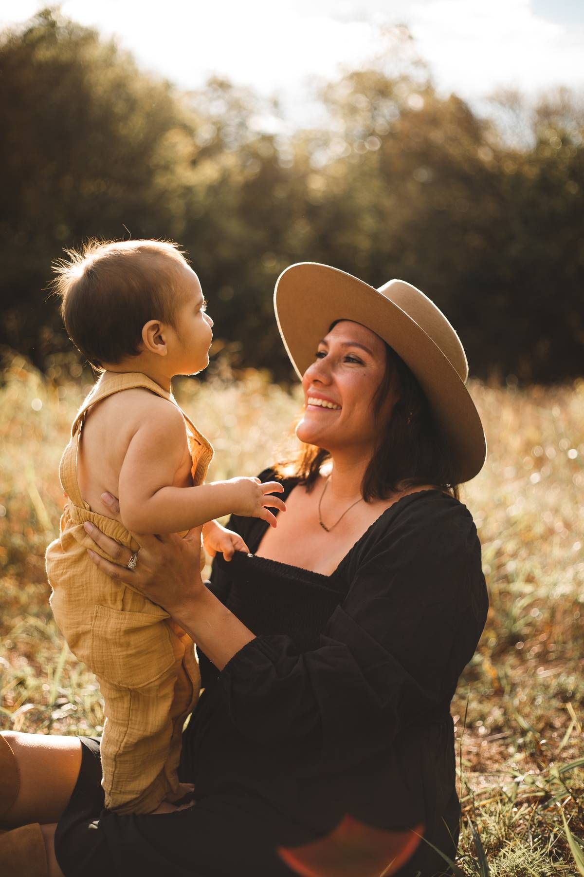 Smiling woman in a wide-brim hat and black top lifts a toddler in a mustard romper while sitting in a golden sunlit field.