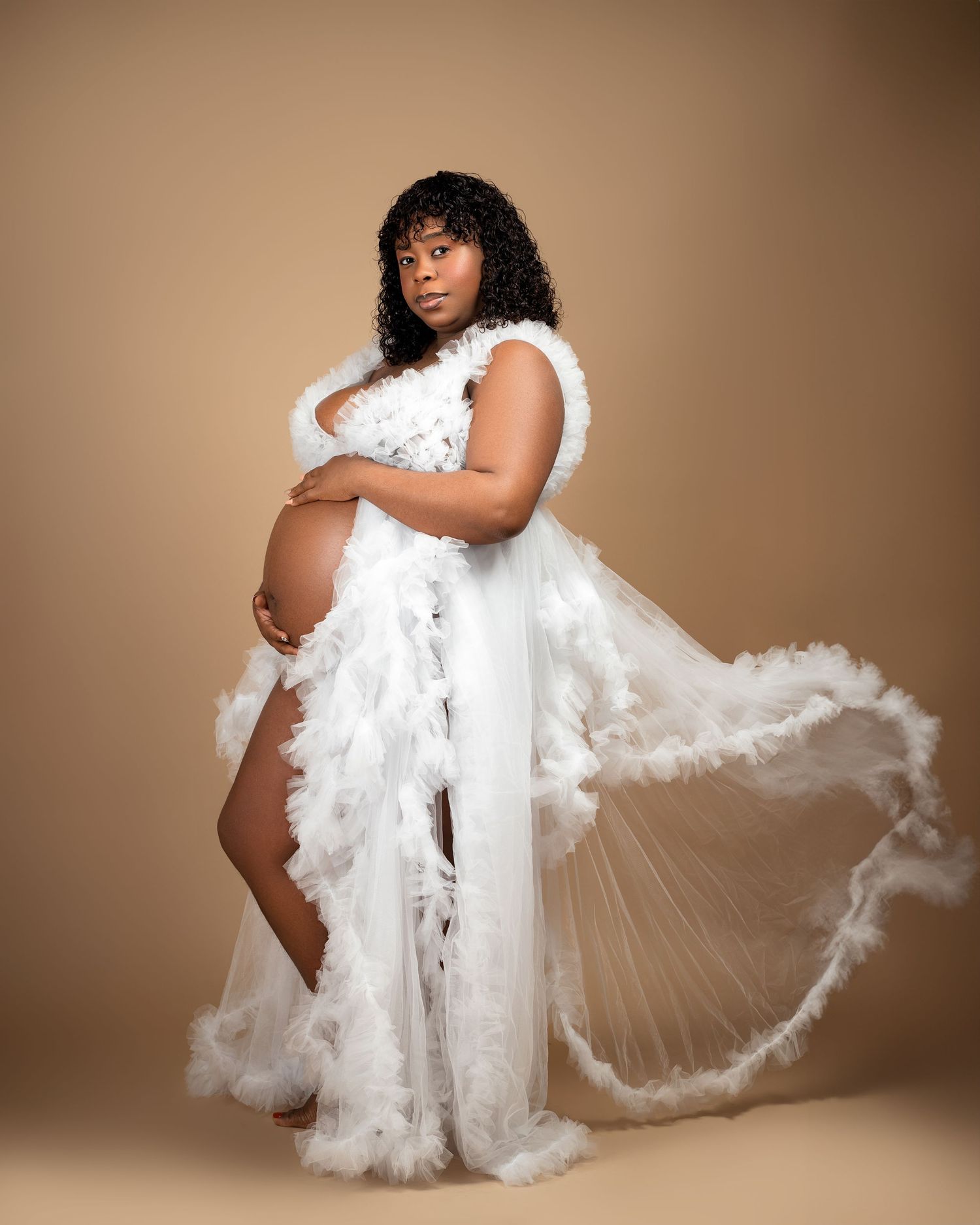 Artistic maternity portrait in flowing white feathered gown against warm backdrop.