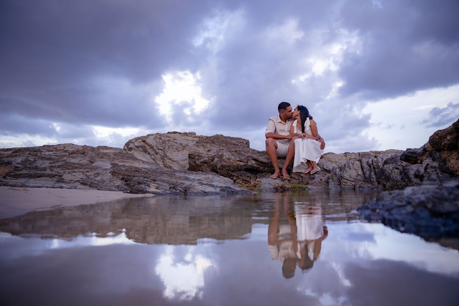 Dramatic cloudy sky reflects in still water at a rocky beach landscape.
