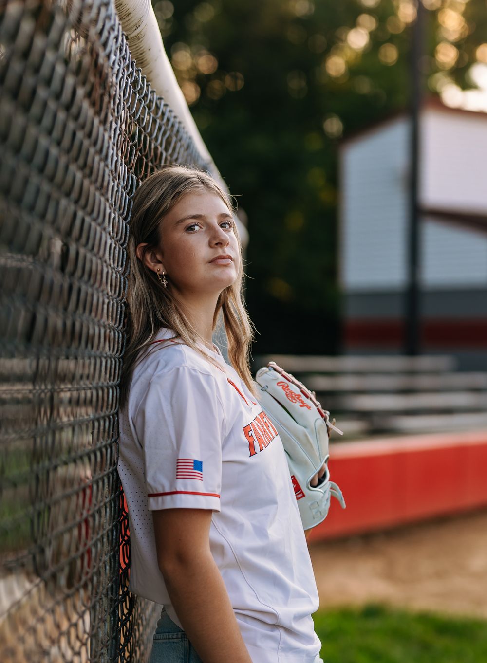 Young female softball player in white jersey leaning against chain-link fence at baseball field during golden hour.