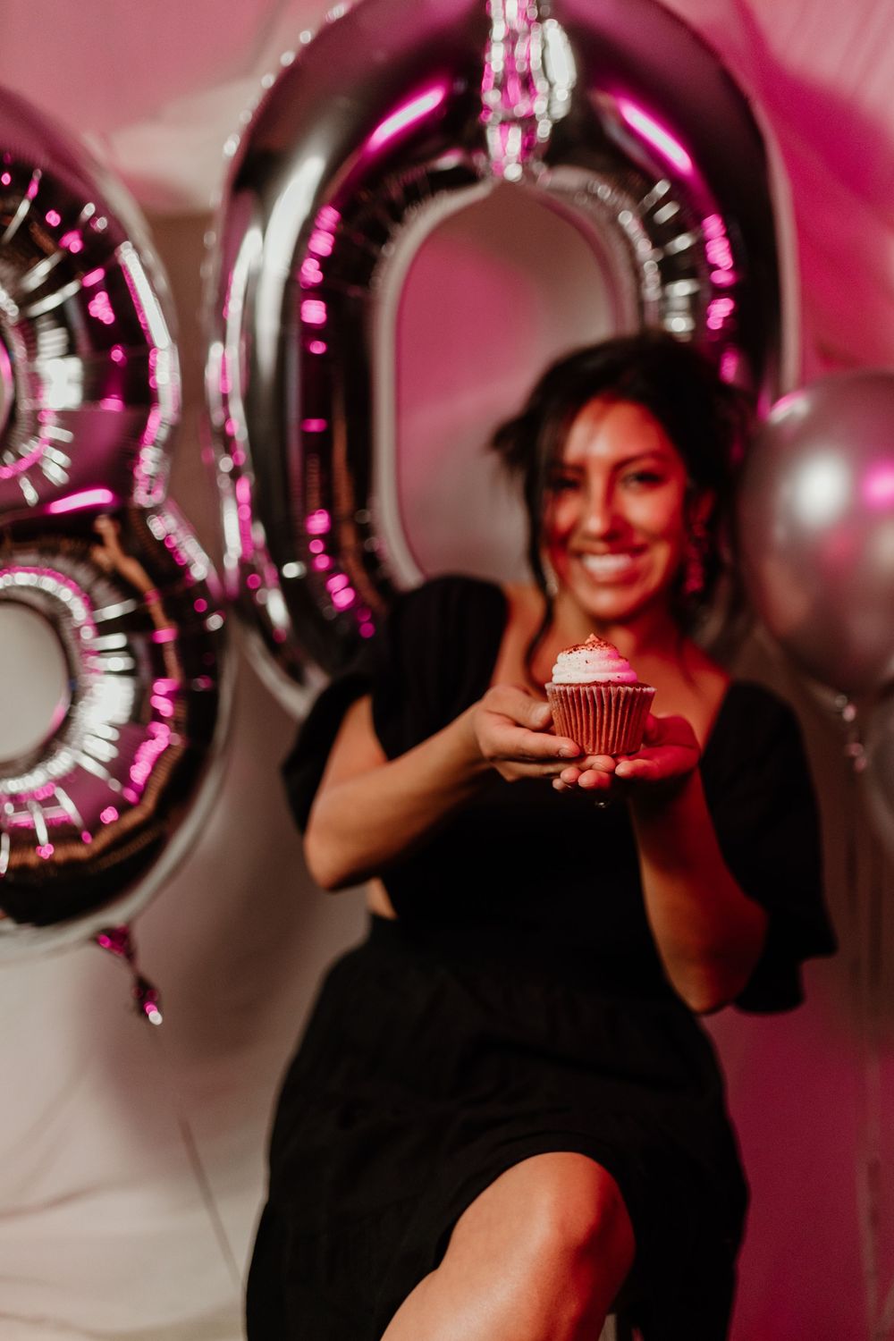 Woman in black dress smiling and holding a birthday cupcake in front of pink number 30 balloons.