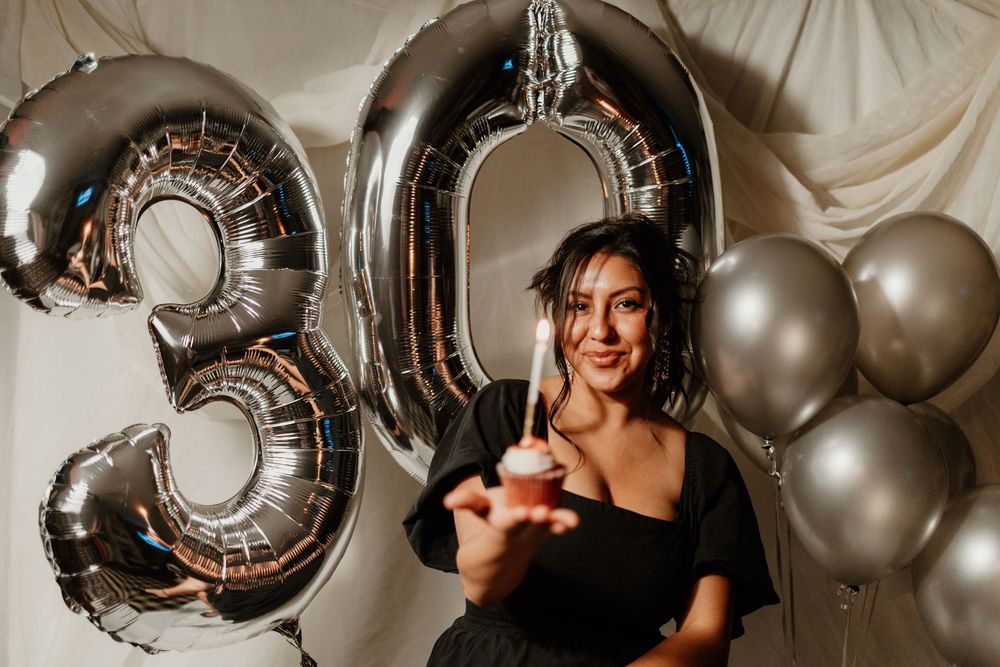 Happy woman in black dress holding cupcake in front of silver number 30 balloons at birthday celebration.