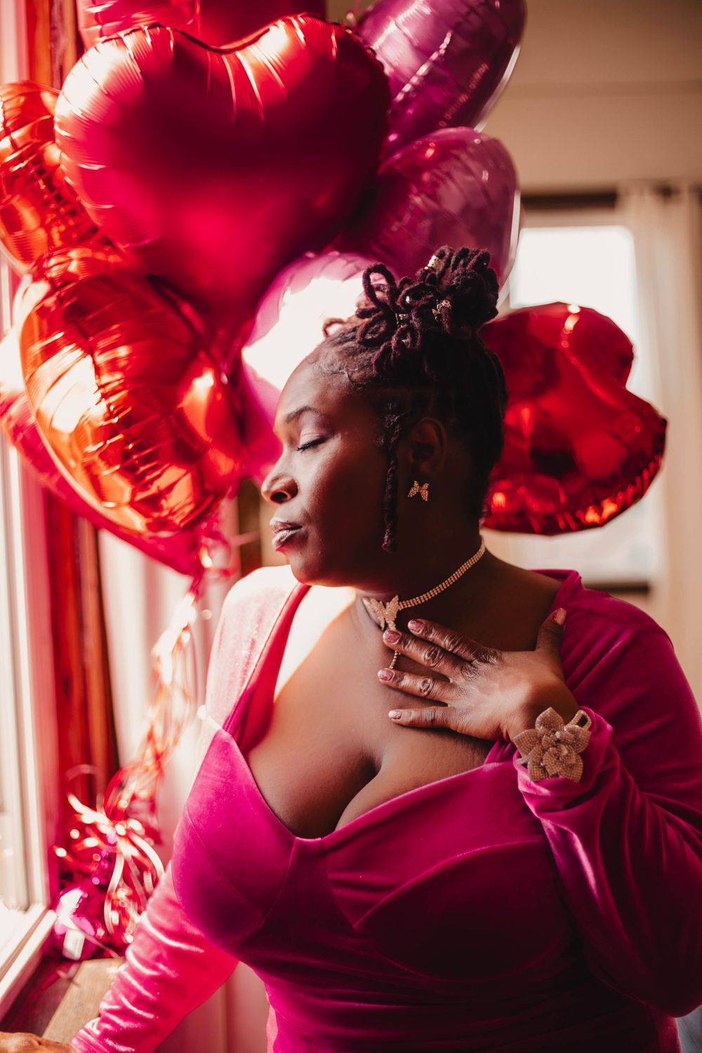 Woman in pink velvet dress poses with red heart-shaped balloons in a Valentine's Day themed portrait.
